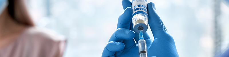 Corona-Schutzimpfung Foto: Großaufnahme von Händen in Einmalhandschuhen, die Impfdosis in Spritze aufziehen. Im Hintergrund ist die Patientin zu sehen.
