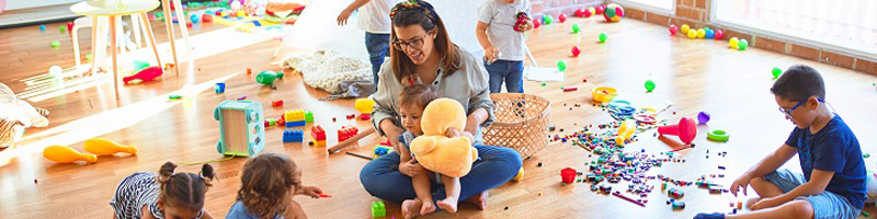 Foto: Betreuerin spielt mit den Kindern am Boden, Möbel und Spielsachen sind zu sehen.