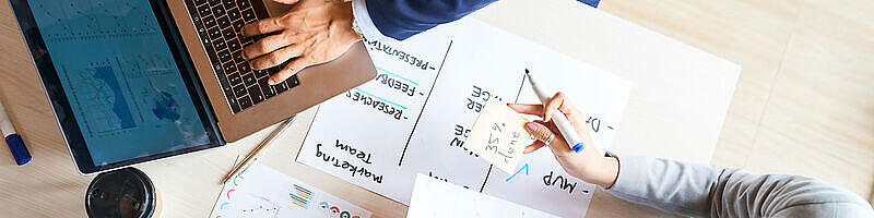 AdobeStock_415332809 Ein Laptop mit darunterliegenden Dokumenten und zwei Hände, die etwas besprechen
