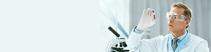 Mann in Laborkleidung mit Schutzbrille betrachtet Proberöhrchen