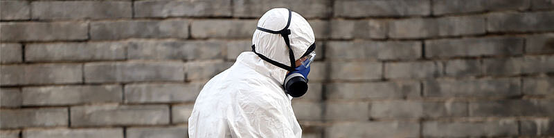 Mensch in Schutzbekleidung mit Atemschutzmaske vor einer Mauer