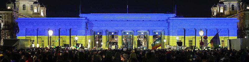 Nächtliches Heldentor in blaugelber Beleuchtung mit demonstrierten Menschen 