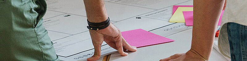 Foto: Tisch mit Flipchartpapier und Postit. 2 Menschen sind teilweise von hinten zu sehen.