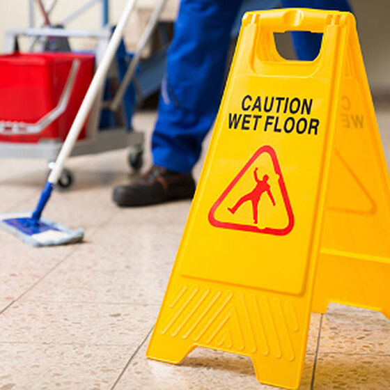 Schild "Caution wet floor". Im Hintergrund wir ein Boden mit Wischmopp gereinigt 