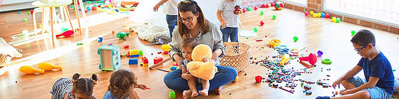 Betreuerin spielt mit den Kindern am Boden, Möbel und Spielsachen sind zu sehen.